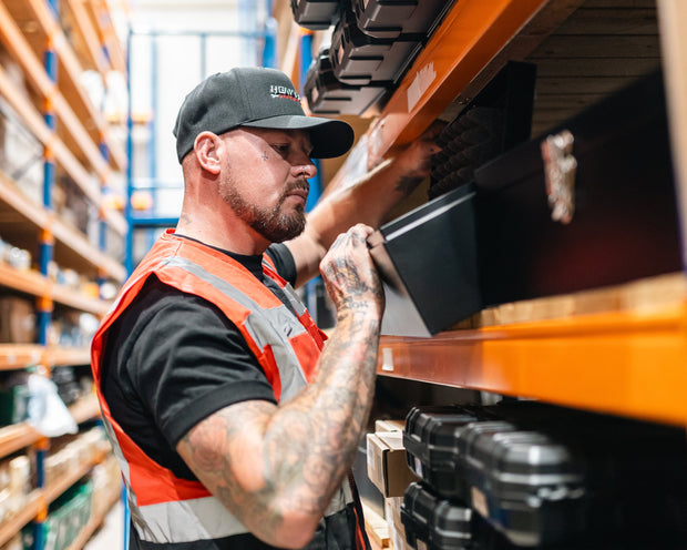 Man in a warehouse wearing a safety vest and cap, working with shelves.